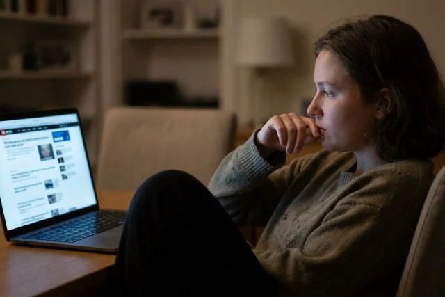 A close-up of a person sitting in a dimly lit room, looking thoughtfully at a glowing laptop screen displaying a news